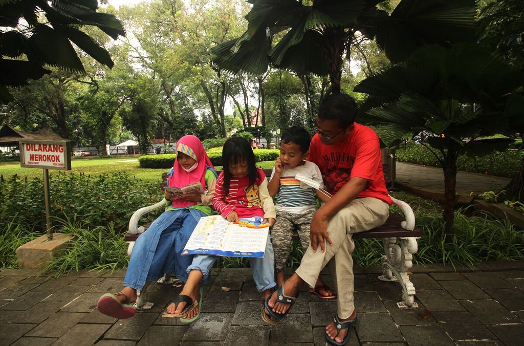 Tiga anak didampingi orangtuanya membaca buku di Taman Suropati, Menteng, DKI Jakarta, Minggu (17/7/2022). Buku itu diambil dari lemari buku Bookhive yang terletak di tengah taman tersebut.