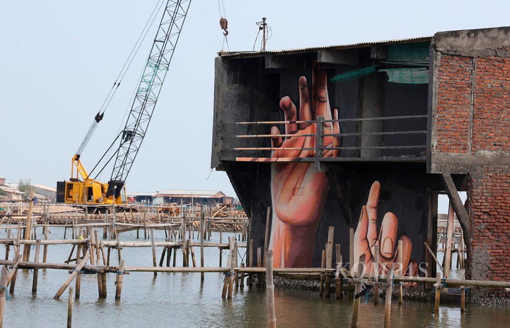 Lukisan lambaian tangan menghiasi rumah yang tenggelam dengan latar belakang proyek pembuatan tanggul laut di Kampung Tambakrejo, Kota Semarang, Jawa Tengah, Kamis (4/5/2023). Pantai utara dengan aktivitas perikanan dan tambaknya saat ini menghadapi banyak persoalan lingkungan. Kerusakan pesisir utara Jawa, antara lain hilangnya kawasan mangrove beralih menjadi permukiman dan lahan industri serta penurunan permukaan tanah, abrasi, hingga perubahan iklim, menjadi penyebabnya.