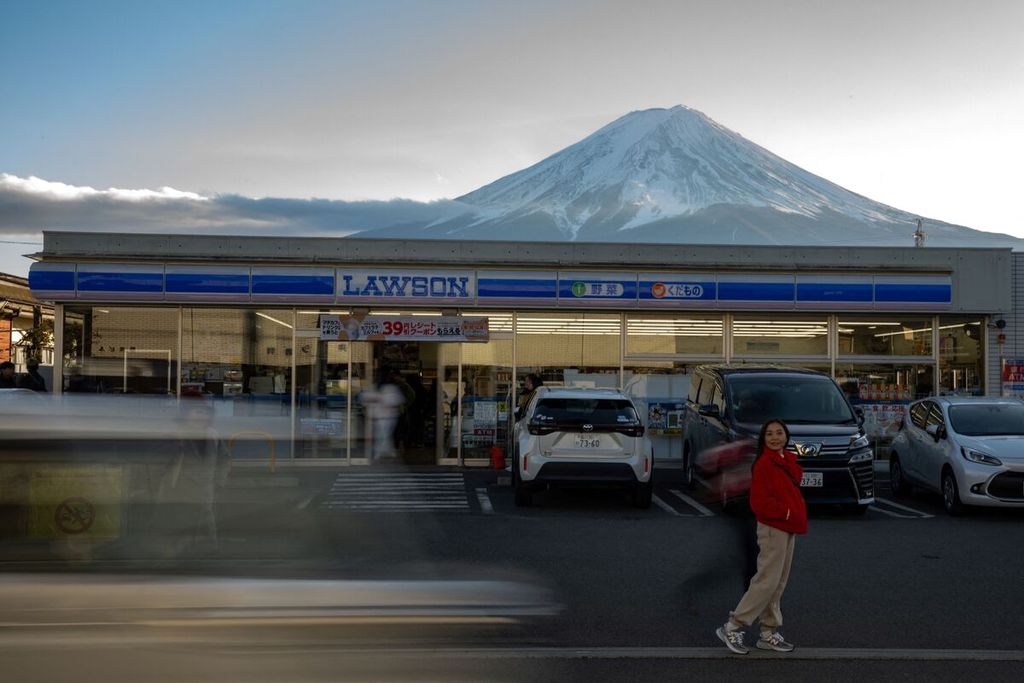 Foto yang diambil pada 1 Januari 2024 ini memperlihatkan seorang turis berpose untuk difoto di depan sebuah toko waralaba dengan latar pemandangan Gunung Fuji di Fujikawaguchiko, Prefektur Yamanashi, Jepang. 