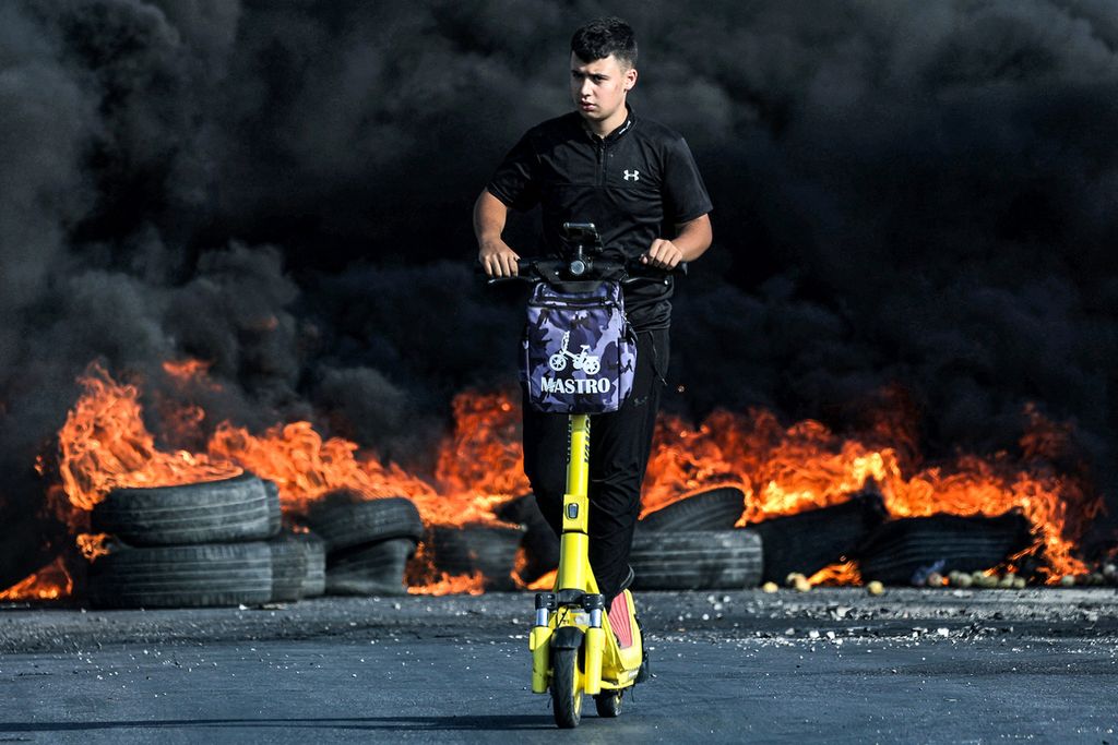 Demonstran Palestina mengendarai skuter melewati ban yang terbakar saat bentrokan dengan tentara Israel di kota Ramallah di Tepi Barat yang diduduki, Minggu (8/10/2023). 