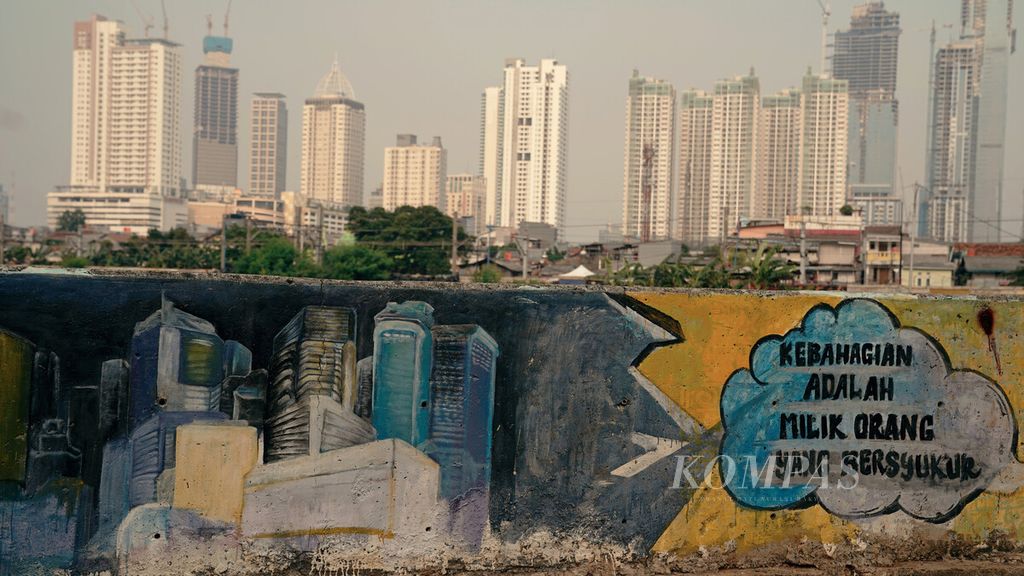 Mural ajakan bersyukur berlatar gedung bertingkat di bantaran Kanal Banjir Barat, Petamburan, Tanah Abang, Jakarta Pusat, Selasa (5/10/2021). Selain migrasi dari desa ke kota, penambahan penduduk perkotaan karena reklasifikasi guna-lahan dari kawasan perdesaan menjadi perkotaan. 