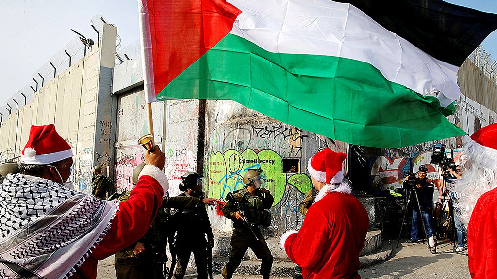 Warga Palestina yang berpakaian ala Sinterklas berdiri di depan tentara Israel dalam unjuk rasa di kota Bethlehem, Tepi Barat, Sabtu (23/12). 