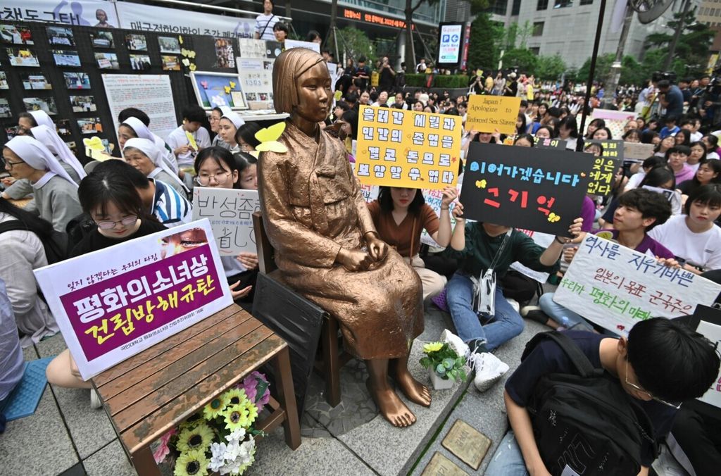 Para demonstran dari Korea Selatan duduk di dekat sebuah patung seorang gadis remaja yang melambangkan ”perempuan penghibur” yang melayani tentara Jepang selama Perang Dunia II pada demonstrasi anti-Jepang dekat Kedutaan Jepang di Seoul pada 24 Juli 2019. 