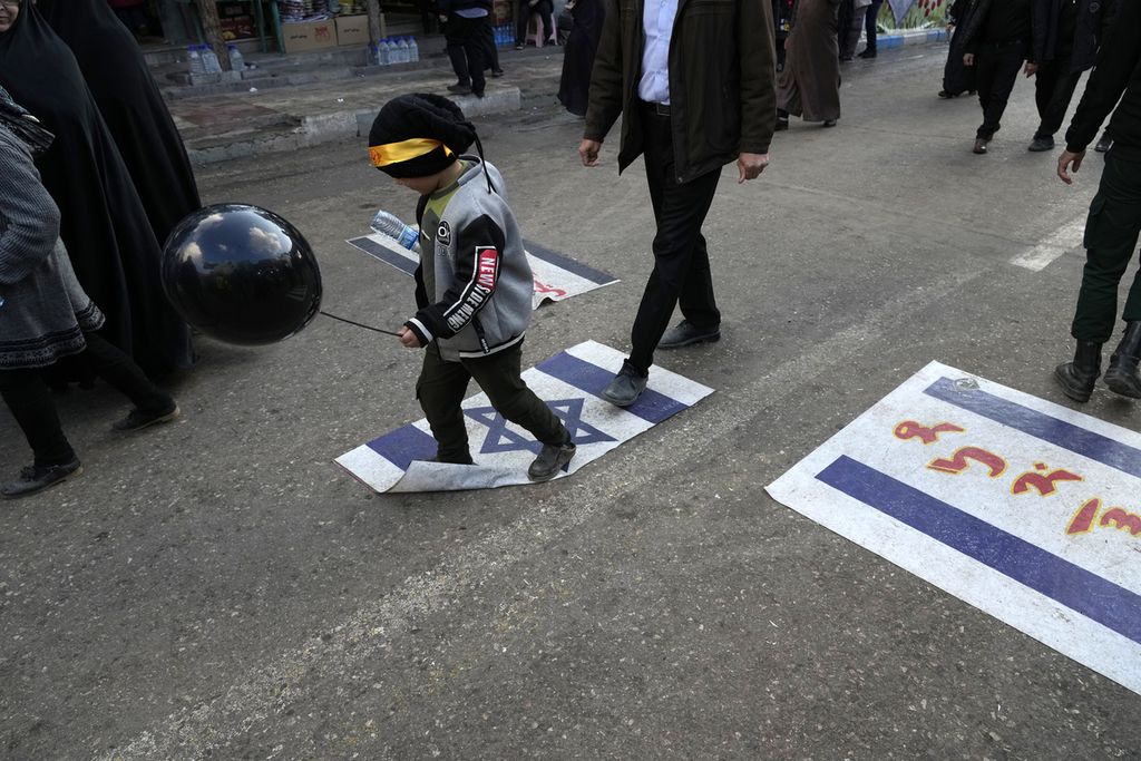 Warga berjalan dengan menginjak bendera Irael di titik peledakan bom di Kerman, Iran, Kamis (4/1/2024). 