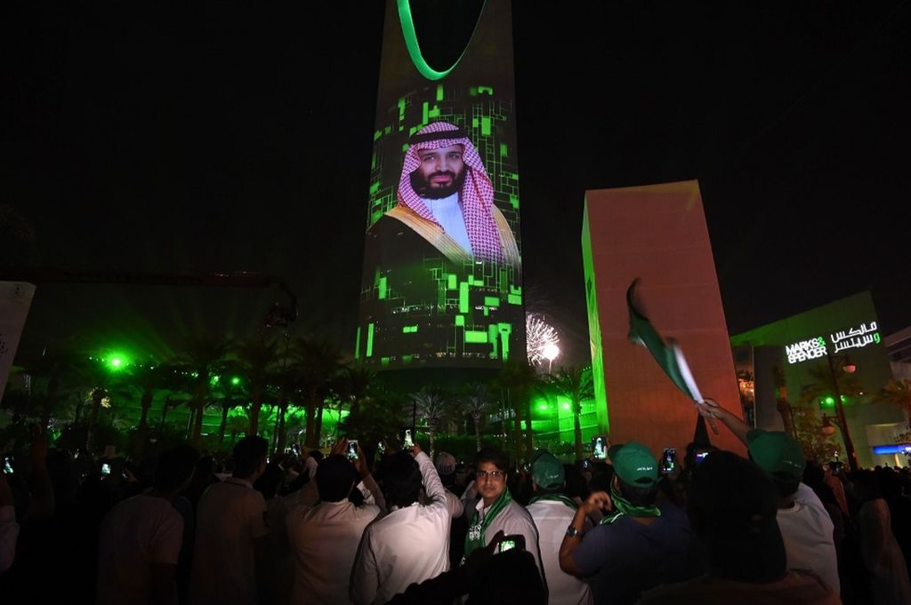 Warga Arab Saudi menyaksikan foto Putra Mahkota Pangeran Mohammed bin Salman, yang dipancarkan ke sebuah gedung pencakar langit di Riyadh, Arab Saudi, Sabtu (23/9), dalam perayaan hari berdirinya Kerajaan Arab Saudi. Pangeran Mohammed bin Salman merupakan tokoh utama yang melahirkan Visi Arab Saudi 2030.