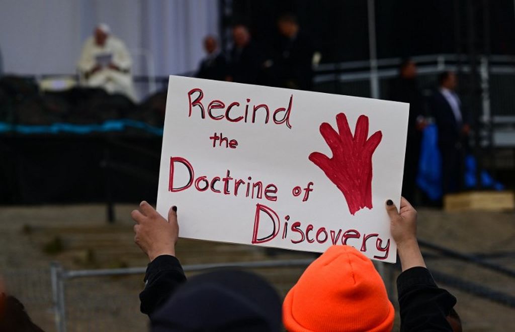 Seseorang yang memegang tanda protes Recind the Doctrine of Discovery (Batalkan Doktrin Penemuan) saat Paus Fransiskus berbicara di panggung di Sekolah Dasar Nakasuk di Iqaluit, Nuvavut, teritori Kanada di Arktik, 29 Juli 2022.