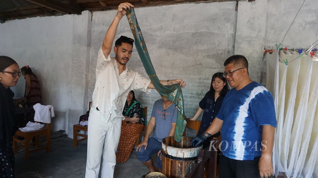 Dino, salah satu peserta tur ke Studio Sejauh Mata Memandang di Pekalongan, Jawa Tengah, Kamis (8/8/2024), melihat hasil pencelupan warna untuk kain yang sudah ia beri motif. Di depannya, Fatah Syaifur Rochman (kanan), pemilik usaha pewarna baju berwarna alam bernama Shibiru, membimbingnya agar mendapat hasil pewarnaan yang bagus.