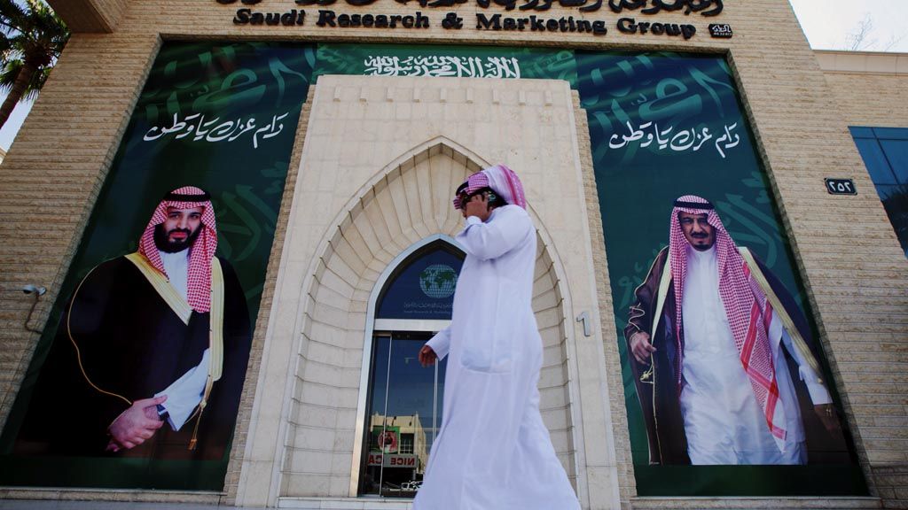 Seorang pria berbicara lewat ponsel, Kamis (9/11), di Riyadh,  sambil berjalan di depan poster yang menampilkan foto Raja Arab Saudi Salman bin Abdulaziz dan Putra Mahkota Pangeran Mohammed bin Salman. Komisi Antikorupsi Arab Saudi, kemarin, membekukan rekening milik mantan Putra Mahkota, Mohammed bin Nayef.