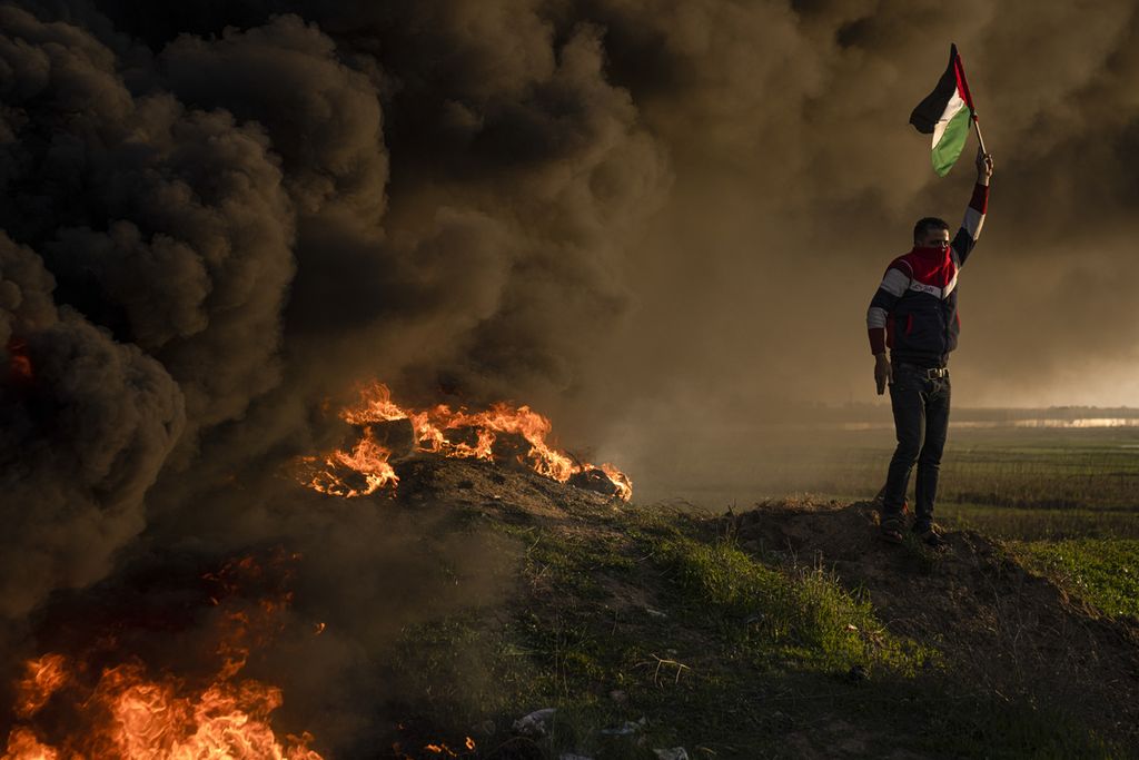 Warga mengibarkan bendera Palestina di tengah kepungan asap hitam yang berasal dari sejumlah ban yang dibakar dalam demonstrasi menentang serangan aparat keamanan Israel ke kamp pengungsi Jenin di Kota Gaza, Kamis (26/1/2023). 