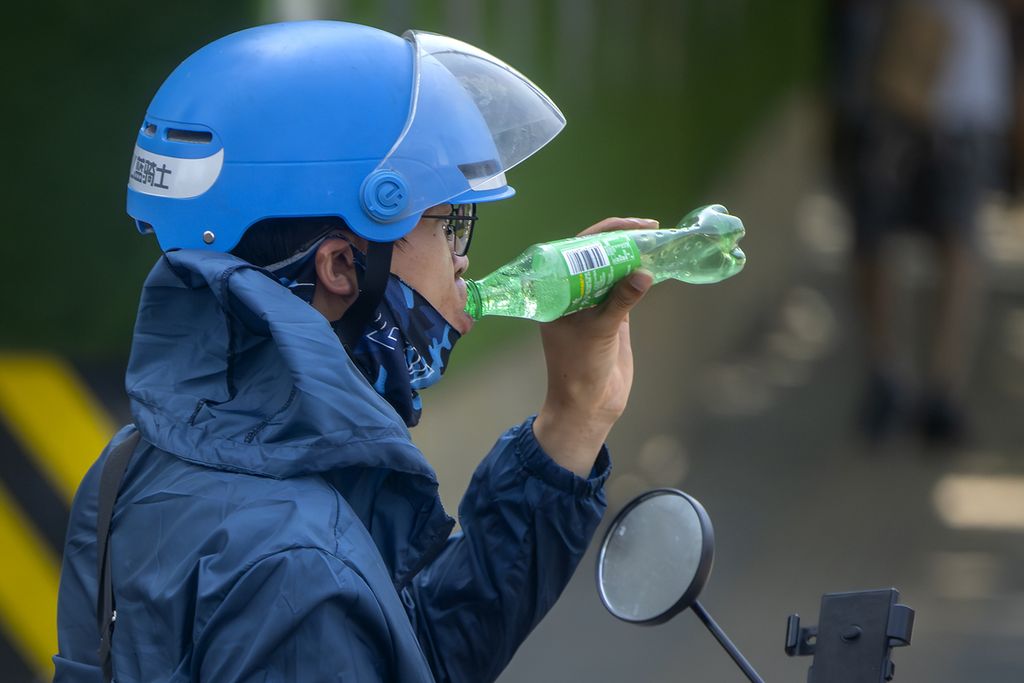 A motorcyclist in Beijing, China, drank water from a bottle while riding under the scorching sun on Saturday (24/6/2023). Chinese weather and health authorities encourage people to reduce outdoor activities and consume water to reduce the risk of dehydration in hot weather.