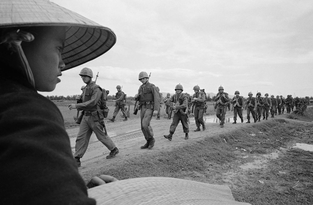 Seorang wanita Vietnam yang mengenakan topi jerami tradisional menyaksikan sekelompok Marinir AS berbaris di sepanjang jalan tanah menuju posisi pertahanan di bukit 327, 15 Maret 1965. Bukit 327 adalah bukit strategis dekat Pangkalan Udara Da Nang. Marinir memperkuat pertahanan pangkalan udara yang penting.