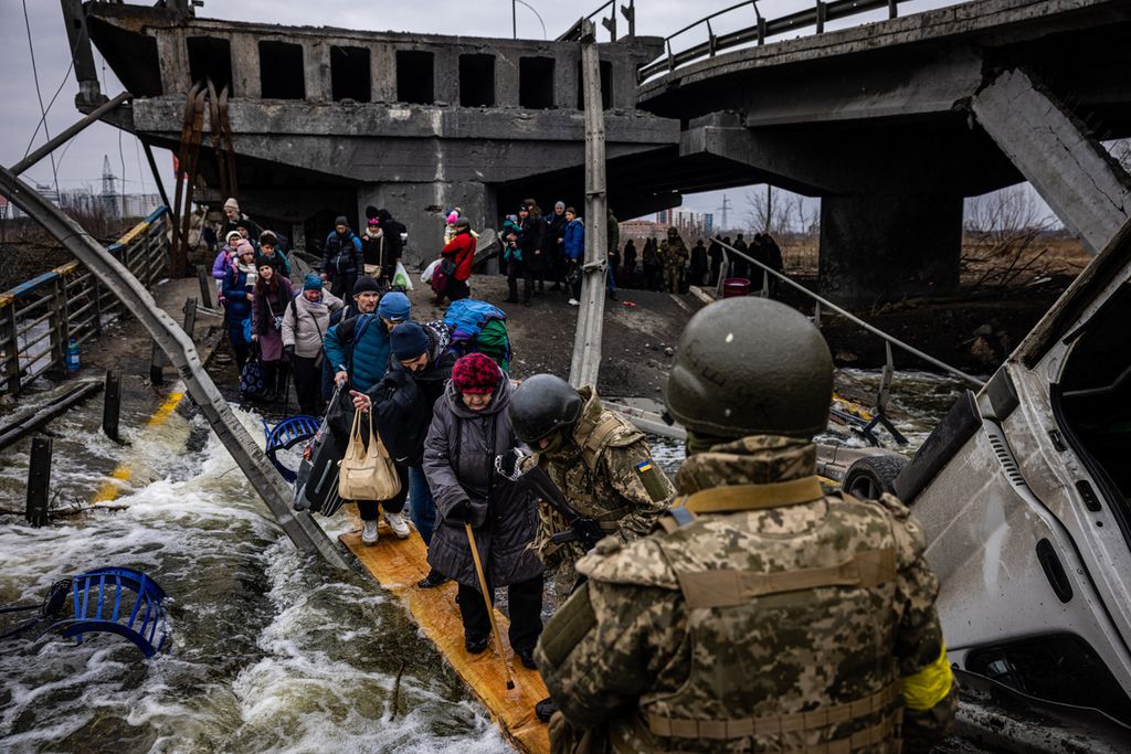 Pengungsi melintasi jembatan yang hancur saat mereka melarikan diri dari kota Irpin, barat laut Kyiv, pada 7 Maret 2022. - Ukraina menolak tawaran Moskwa untuk mendirikan koridor kemanusiaan dari beberapa kota yang dibombardir pada hari Senin setelah muncul beberapa rute yang akan membawa pengungsi ke Rusia atau Belarusia. 