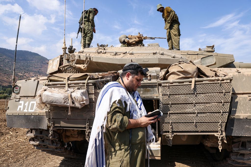 Seorang tentara Israel berdoa sambil berdiri di depan tank Merkava di pinggiran kota utara Kiryat Shmona dekat perbatasan dengan Lebanon, Minggu (8/10/2023). 