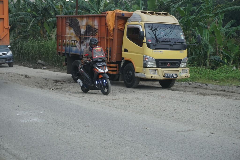 Sepeda motor berjalan berdampingan dengan truk di jalan berlubang. Truk-truk pembawa hasil tambang dari Parung Panjang, Bogor, Jawa Barat, yang bermuatan berlebih diduga menjadi penyebab jalan rusak.