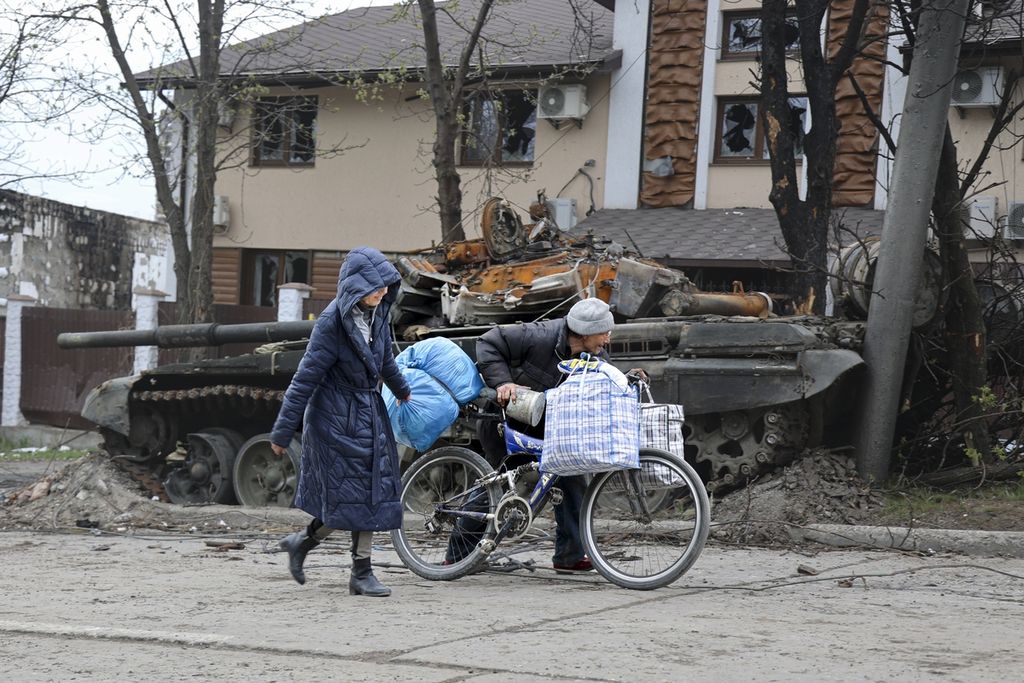 Warga berjalan melalui sebuah tank yang rusak akibat pertempuran antara kelompok separatis yang didukung militer Rusia dan militer Ukraina di Mauripol, Ukraina, Selasa (19/4/2022). Mokswa dilaporkan tengah berupaya keras mengambil alih Mauripol. 
