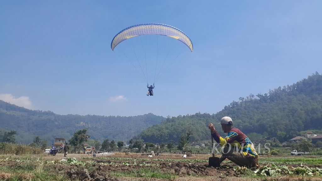 Seorang wisatawan domestik terbang tandem dari atas Gunung Banyak dan bersiap mendarat di Lapangan Songgo Maruto, Songgokerto, Kota Batu, Jawa Timur, Rabu (9/8/2023). Tampak di bawahnya seorang petani beraktivitas di lahan.
