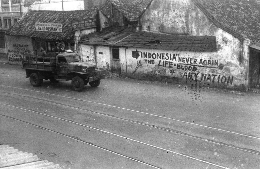 Jakarta, 11 Desember 1945. Truk tentara sekutu melintasi dinding-dinding bertuliskan semboyan perjuangan Indonesia, diduga di kawasan Senen, Jakarta Pusat. 