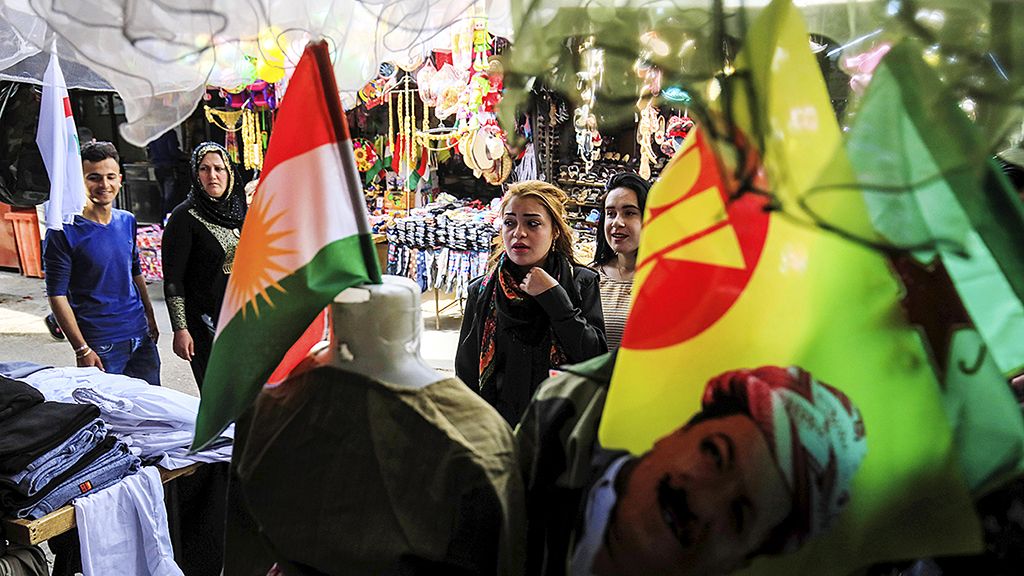 Perempuan Kurdi, Selasa (20/3), berada  di kios sebuah pasar di kota Qamishli,  Suriah timur laut, menjelang perayaan tahun baru  Noruz. Bendera Kurdi dan mantan Presiden Kurdistan Irak Massud Barzani tampak di latar depan. 