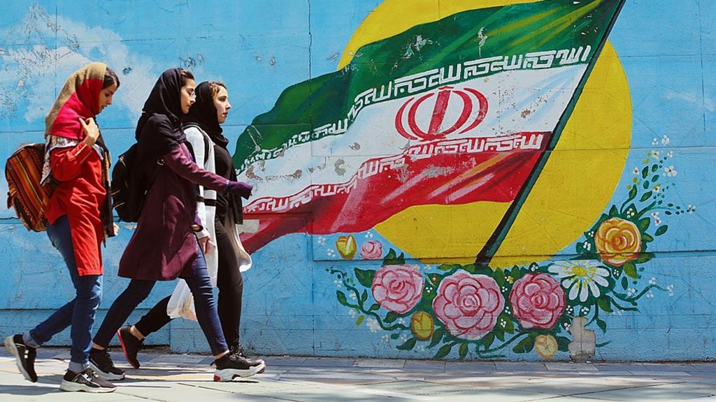 Anak-anak perempuan Iran berjalan melewati sebuah mural bergambar bendera nasional Iran di Teheran, Iran, Selasa (23/4/2019). AS telah mengumumkan penghentian keringanan dari sanksi pada sejumlah negara yang mengimpor minyak Iran.