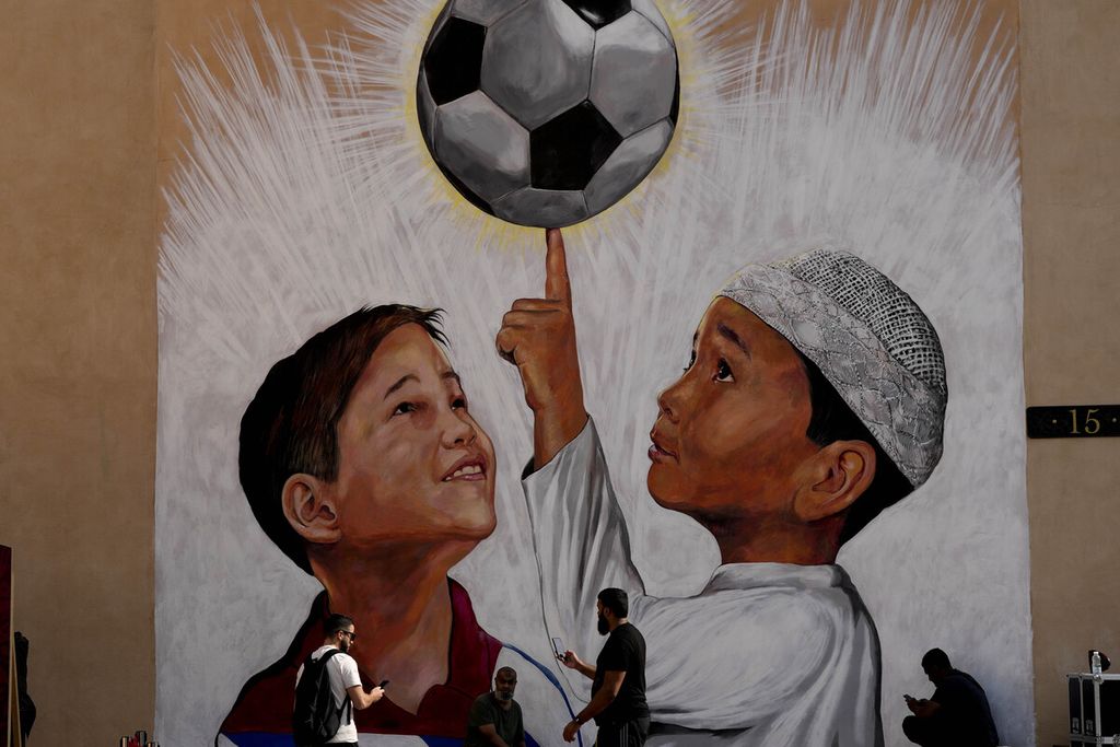 Beberapa warga berjalan di depan sebuah mural yang menggambarkan dua anak, salah satunya berupaya menjaga keseimbangan bola dengan jari telunjuknya, di Doha, Qatar, 7 Desember 2022. Qatar menggunakan sepak bola dan ajang olahraga sebagai bagian dari upaya mengubah imaji negaranya dalam pandangan global. 