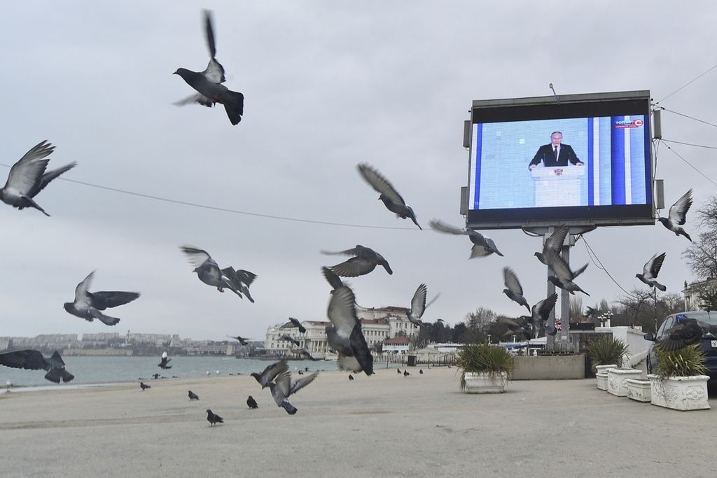 Burung-burung merpati beterbangan di pantai dengan layar besar yang sedang menayangkan pidato Presiden Rusia Vladimir Putin di Sevastopol, Crimea, Selasa (21/2/2023). 
