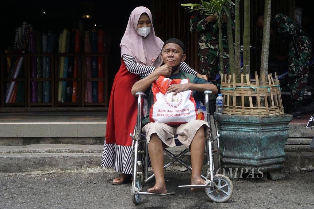 Rubianti (59) berdiri memegang kursi roda tempat suaminya, Pardi Subono (60), duduk dengan kondisi stroke di Pasar Klandasan Balikpapan, Kalimantan Timur, Selasa (25/10/2022). Sebelumnya, mereka berjejalan di antara pengunjung pasar untuk melihat Presiden Joko Widodo yang berkunjung.