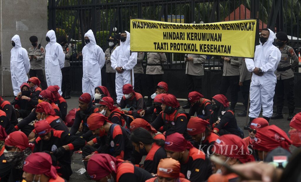 Police wearing personal protective equipment (PPE) unfurls a banner appealing to health protocols when thousands of workers demonstrate in front of the DPR Building, Senayan, Jakarta, Monday (7/2/2022). The mass of workers from the Federation of Indonesian Metal Workers Union (FSPMI) and other elements again demonstrated by demanding to reject the Job Creation Law and ask for a revision of the minimum wage in areas other than DKI Jakarta.