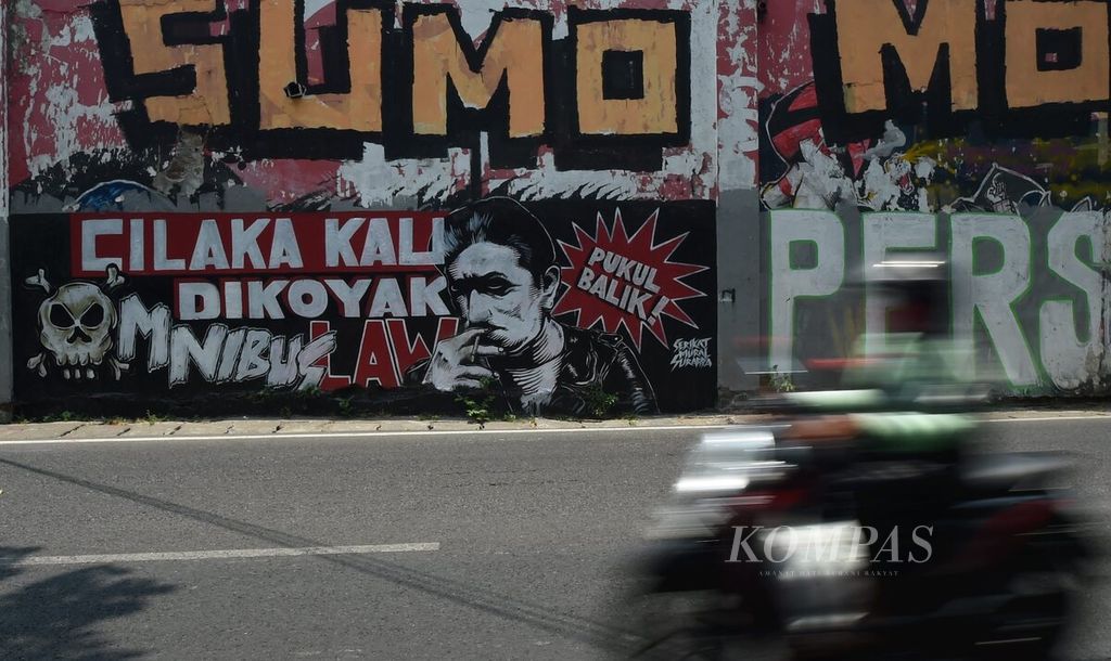 Pengendara melintasi mural tema tolak UU Cipta Kerja yang dibuat oleh Serikat Mural Surabaya di Jalan Dinoyo, Surabaya, Rabu (7/10/2020). Gelombang penolakan terhadap UU Cipta Kerja terus dilakukan oleh banyak pihak. Selain unjuk rasa, penolakan juga dilakukan melalui berbagai media salah satunya mural.