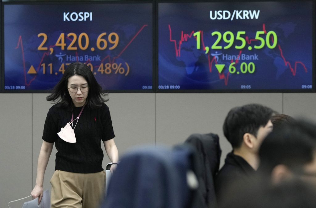 Seorang pialang tengah berjalan melintas di depan layar yang memperlihatkan pergerakan saham di Korea Composite Stock Price Index (KOSPI), dan nilai tukar dollar AS terhadap won Korea Selatan pada Selasa (28/3/2023).