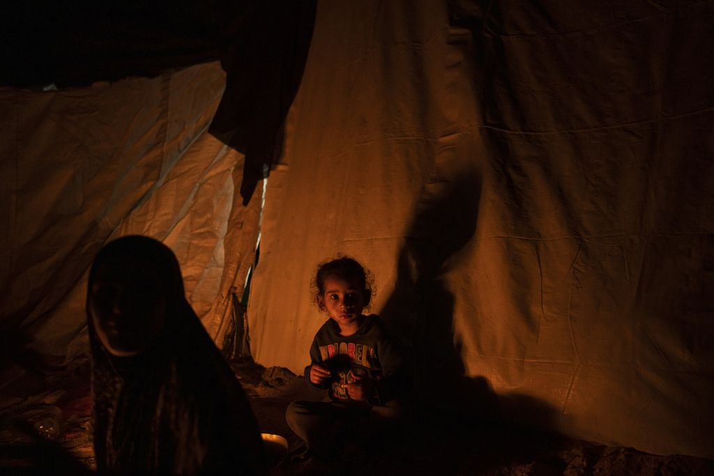 Seorang anak dan orangtuanya tinggal di dalam sebuah tenda yang diterangi cahaya api di kamp pengungsian Muwasi, Minggu (31/12/2023). Malam pergantian tahun kali ini dilalui warga Gaza dalam suasana perang. 