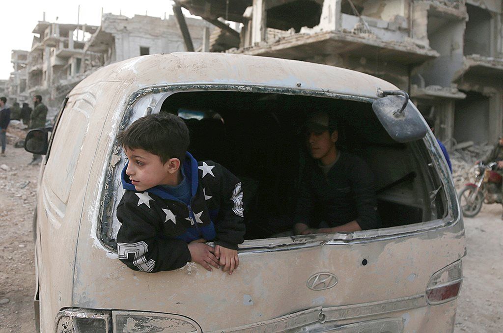 Anak-anak Suriah terlihat di belakang kendaraan yang hancur ketika warga tengah bersiap keluar dari Arbin, salah satu wilayah tersisa yang masih dikuasai pemberontak. Negosiasi antara pemberontak dan pemerintah Suriah menghasilkan kesepakatan, pemberontak dan keluarga mereka keluar dari Ghouta timur. Ribuan warga sipil di Ghouta timur turut mengungsi. 