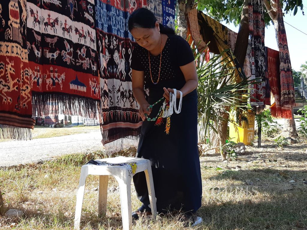 Salah seorang ibu tengah memajang beragam gelang tradisional di dekat kain ikat Sumba yang digantung di halaman rumahnya di Desa Raja Prailiu, Kecamatan Kambera, Sumba Timur, Rabu (24/5/2023) pagi.