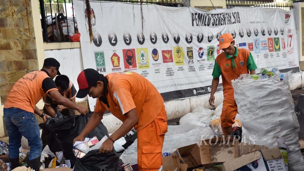 Petugas kebersihan mengangkut sampah dengan latar belakang spanduk sosialisasi partai peserta pemilu serentak 2019 yang terpasang di pagar Kantor Komisi Pemilihan Umum (KPU) Pusat, Jakarta, Rabu (20/3/2019).