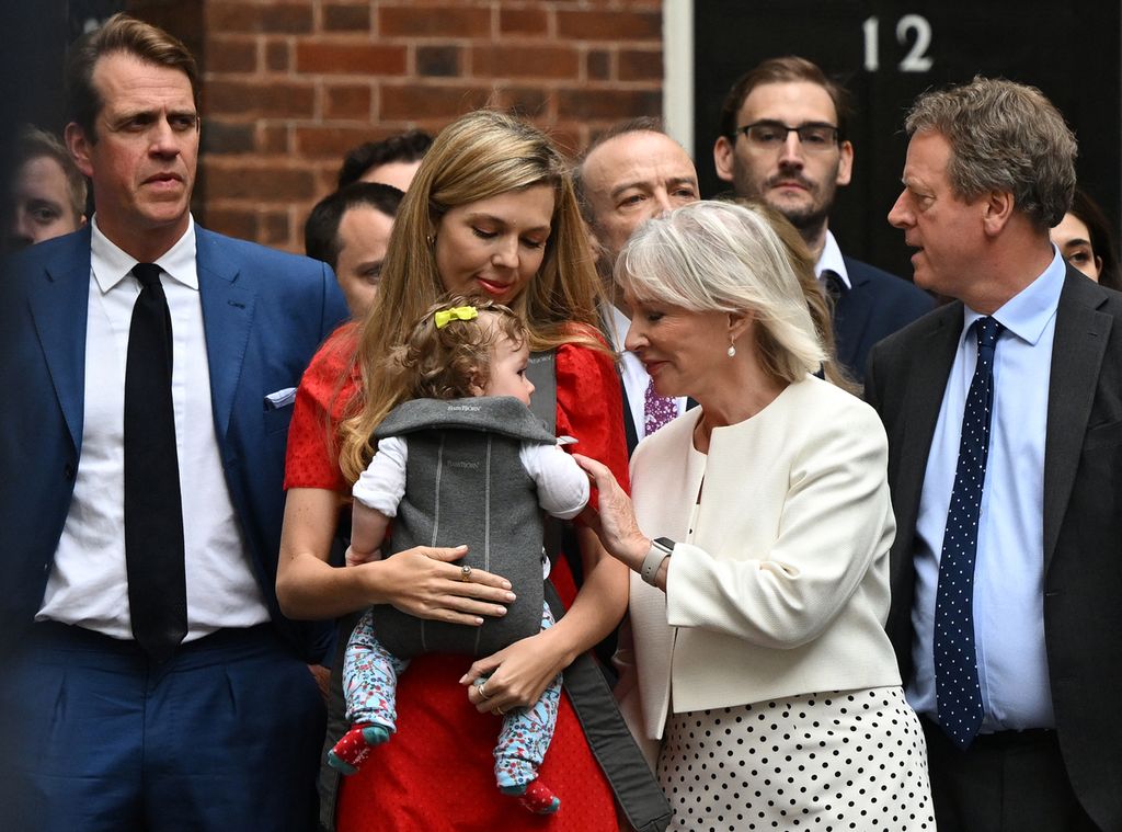 Carrie Johnson, istri Perdana Menteri Inggris Boris Johnson (bergaun merah, tengah), menggendong putrinya ditemani sejumlah kolega dan anggota kabinet, mendengarkan pengumuman pengunduran diri sang suami dari jabatannya, Kamis (7/7/2022). 