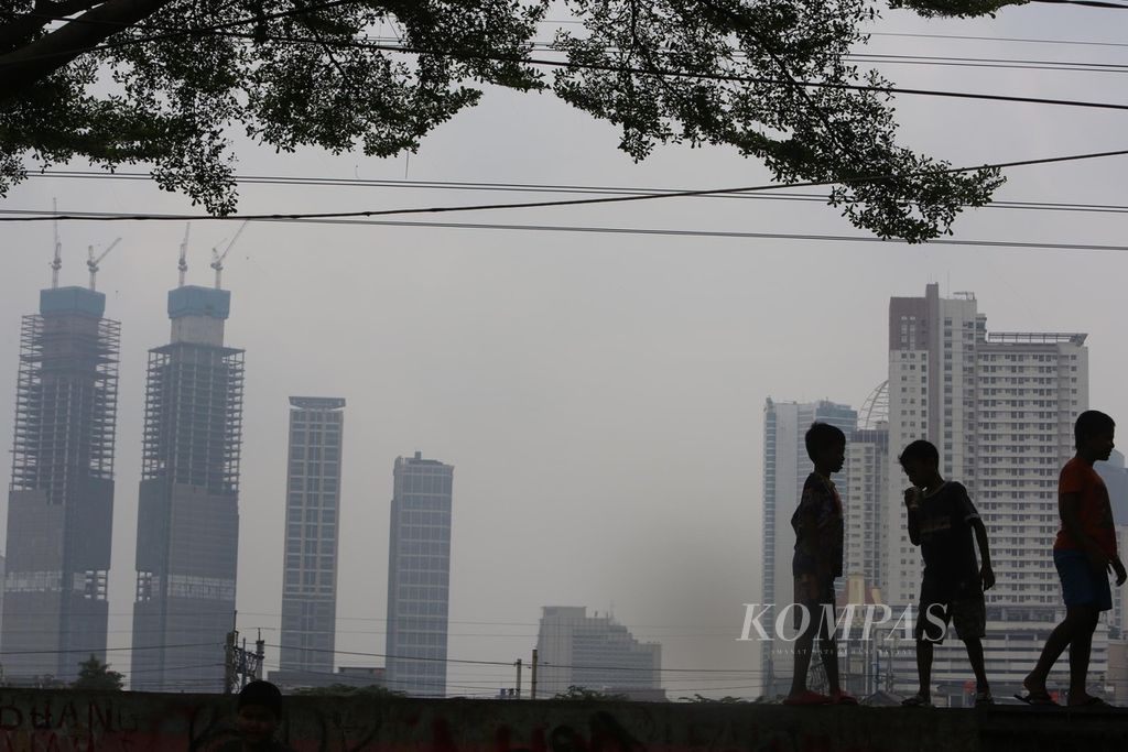 Anak-anak bermain di atas tanggul Sungai Ciliwung dengan latar belakang deretan gedung bertingkat Kota Jakarta yang diselimuti kabut asap polusi, Rabu (15/6/2022). Menurut data situs penyedia peta polusi daring harian kota-kota besar di dunia, AirVisual, pada pukul 11.00 kemarin, nilai Indeks Kualitas Udara (AQI) kota Jakarta adalah 190 atau masuk dalam kategori tidak sehat. Sementara kandungan partikel halus di udara yang ukurannya 2,5 mikron atau lebih kecil (PM2,5) sebesar 131 mikrogram per meter kubik, jauh di ambang batas maksimal 5 mikrogram per meter kubik.
