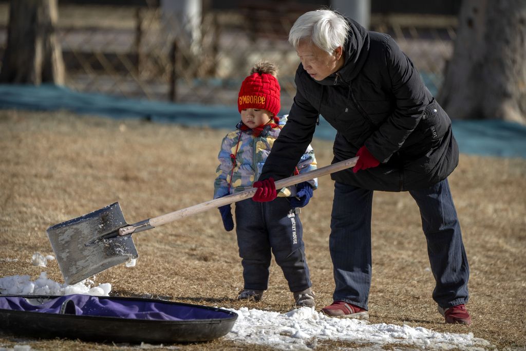 Seorang anak memperhatikan saat seorang pria mengambil salju dengan sekop di sebuah taman di Beijing, China, 17 Januari 2023. China mengumumkan penurunan populasi untuk pertama kalinya dalam beberapa dekade. 