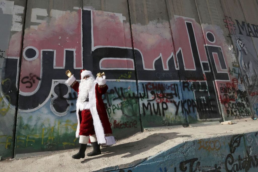 Warga berpakaian Santa Claus berpose di depan grafiti bertuliskan Palestina dalam aksara Arab di dinding pemisah Israel di Bethlehem, Tepi Barat, Sabtu (2/12/2023). 