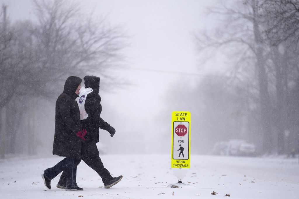 Dua orang berjalan melintasi sebuah penyeberangan di jalan yang tertutup salju di St Louis, Missouri, Kamis (22/12/2022). 