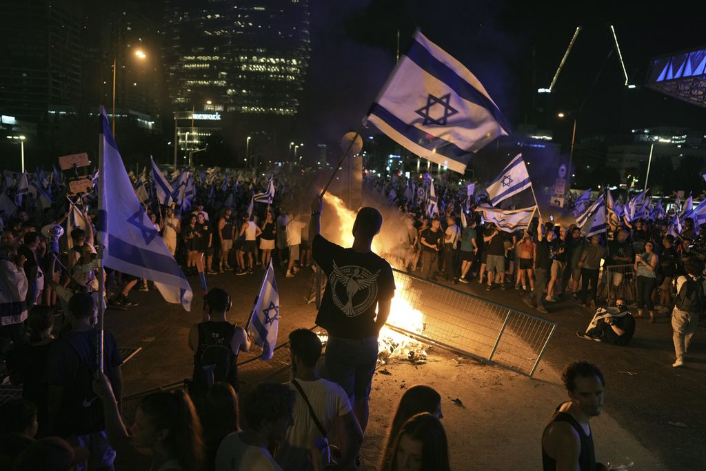 Para pengunjuk rasa membakar ban bekas sambil membentangkan bendera Israel di Tel Aviv, Senin (24/7/2023) malam, pascapengesahan UU Reformasi Hukum oleh parlemen. 