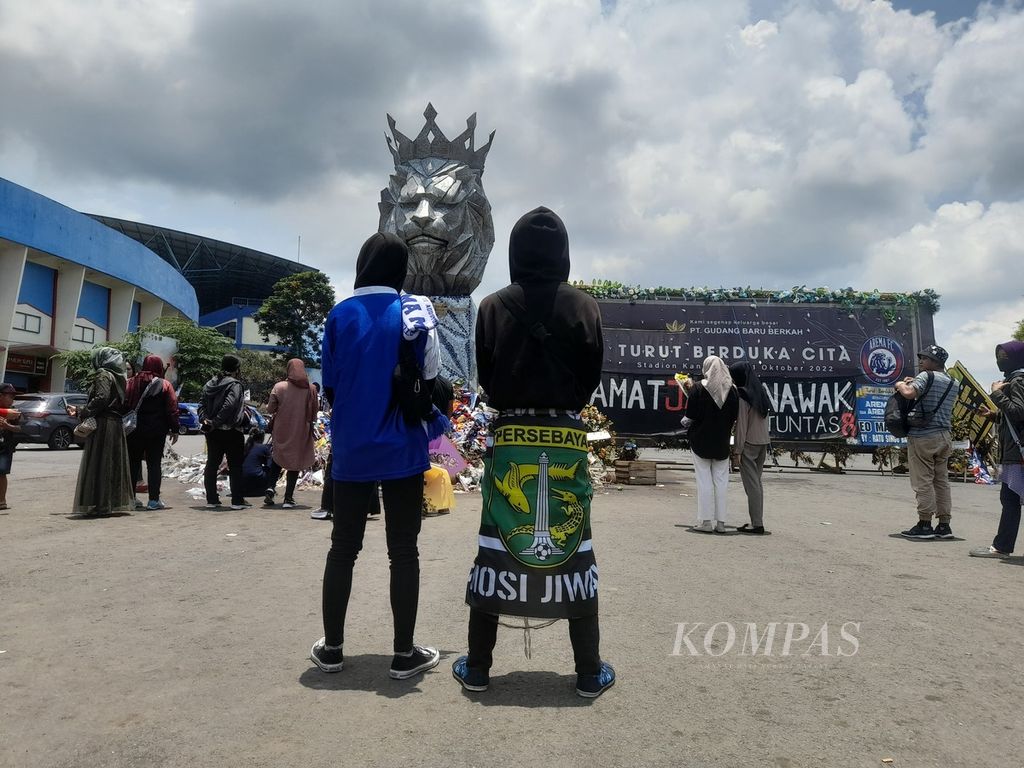 Dua orang pendukung sepakbola yang mengenakan atribut Arema (biru, kiri) dan Persebaya (hijau, kanan) tampak berdiri di depan patung singa di halaman Stadion Kanjuruhan di Kepanjen, Kabupaten Malang, Jawa Timur, sebagaimana terlihat Minggu (23/10/2022) atau tiga pekan seusai Tragedi Kanjuruhan.