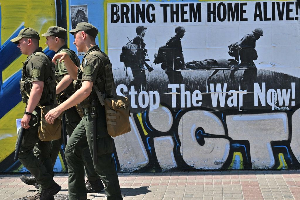 Prajurit Ukraina berjalan melewati sebuah poster yang berbunyi Bring them home alive! Stop the war now! (Bawa mereka pulang hidup-hidup! Stop perang sekarang juga!) di pusat kota Kyiv, Ukraina, Selasa (28/6/2022).