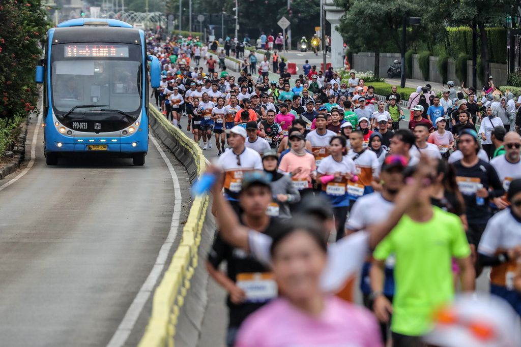 Pelari LPS Monas Half Marathon berlari di samping bus Transjakarta di Jalan Jenderal Sudirman, Jakarta, Minggu (2/7/2023). 