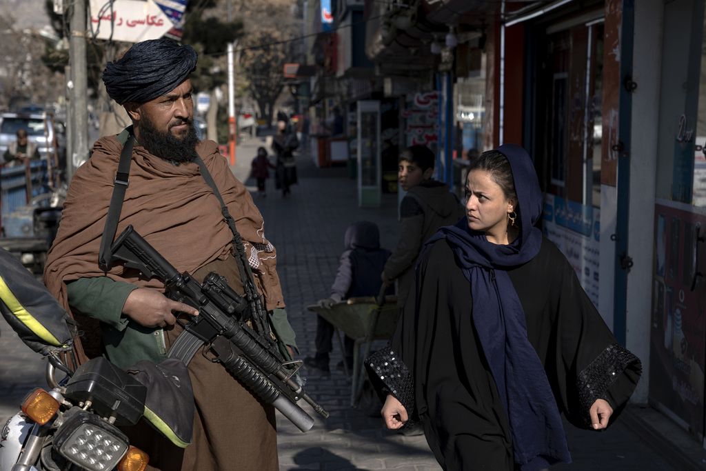 Seorang petempur Taliban berjaga-jaga dengan senapannya, sementara seorang perempuan berjalan melintas di sebelahnya di Kabul, Afghanistan, 26 Desember 2022. 