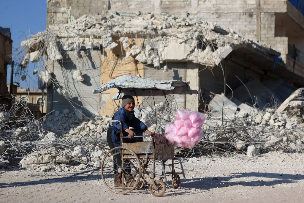 Seorang anak Suriah menjual gula-gula kapas tersenyum saat berdiri di depan reruntuhan bangunan akibat gempa di Atarib, Provinsi Aleppo, yang dikuasai kelompok pemberontak, 14 Februari 2023. 