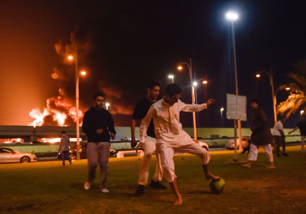 Sejumlah remaja bermain bola di Jeddah, Arab Saudi, tak jauh dari lokasi ledakan fasilitas penyimpanan minyak milik Saudi Aramco yang terbakar akibat serangan rudal dari milisi Yaman, Jumat (26/3/2022) malam. 