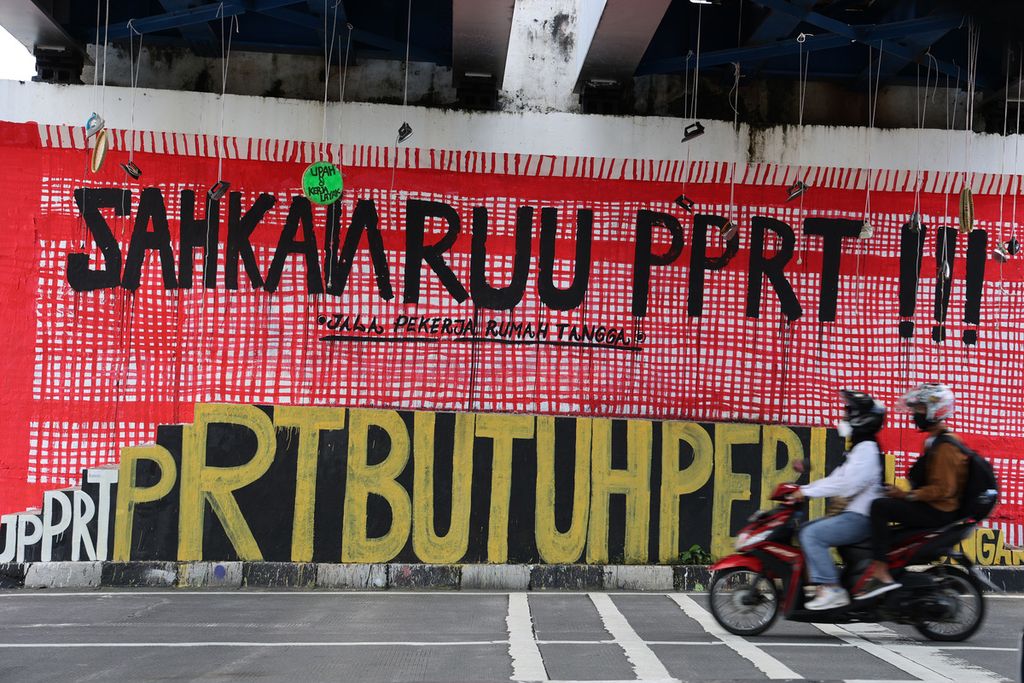 Pengguna sepeda motor melintas di depan mural berisi kritik tentang pembahasan RUU Perlindungan Pekerja Rumah Tangga yang tidak kunjung selesai di Jembatan Kewek, Yogyakarta, Selasa (28/12/2021). 