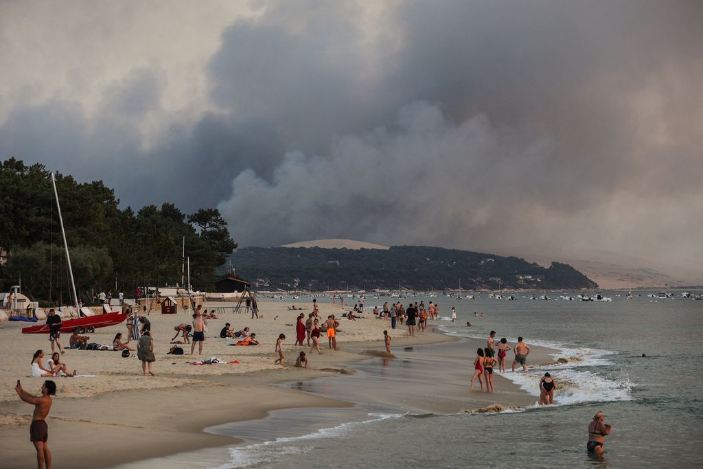Foto yang diambil pada 18 Juli 2022 ini memperlihatkan orang-orang berenang di Pantai Moulleau saat asap mengepul dari kebakaran hutan di La Teste-de-Buch, Perancis, terlihat dari Arcachon, di depan bukit pasir Pilat. Dalam panas terik, dengan suhu lebih dari 40 derajat celsius, sekitar 8.000 orang harus meninggalkandengan "cara pencegahan" menurut prefekturMiquelots dan Pyla-sur-Mer, Distrik La Teste-de-Buch, kota dengan 28.000 penduduk di mana 4.300 hektar hutan menjadi asap. 