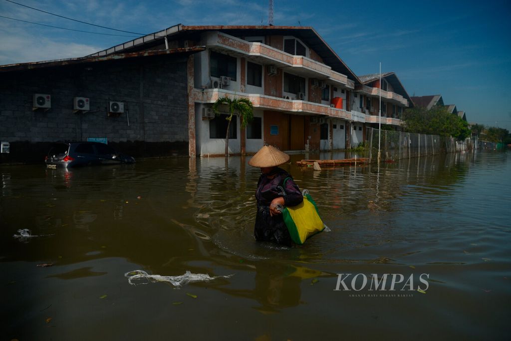 Warga melintas di antara genangan banjir pasang air laut dengan latar belakang pabrik yang berhenti beroperasi di kawasan berikat Pelabuhan Tanjung Emas, Kota Semarang, Jawa Tengah, Jumat (27/5/2022). 