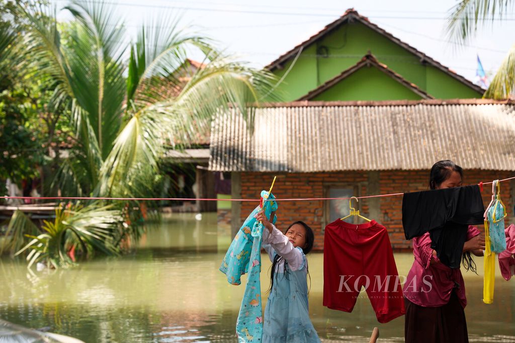 Anak-anak membantu ibunya menjemur pakaian dengan latar belakang rumah yang terendam banjir di Kecamatan Karanganyar, Kabupaten Demak, Jawa Tengah, Selasa (19/3/2024).