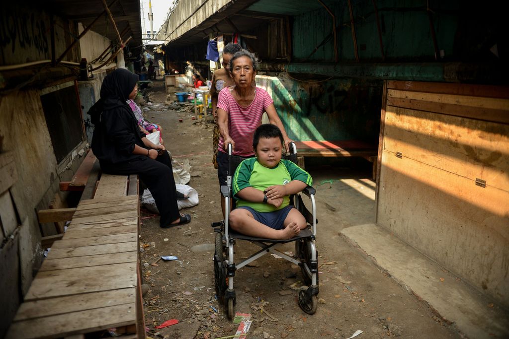 Seorang anak dibantu orangtuanya menggunakan kursi roda menuju sekolah yang ada di permukiman di bawah Jalan Tol Cawang-Tomang-Pluit, Jelambar, Jakarta Barat, Senin (19/6/2023). 